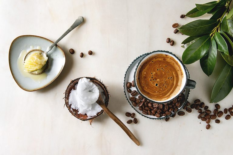 Bulletproof coffee. Keto diet coffee in blue ceramic cup with organic ghee butter and coconut cold press oil in spoons with beans and green branch over white marble background. Flat lay, space